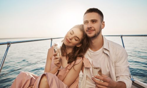 Outdoor portrait of adult happy male and female in love, celebrating their engagement on yacht, holding glass of champagne and hugging. Boyfriend just told about things he likes in her. Emotions and vacation concept.